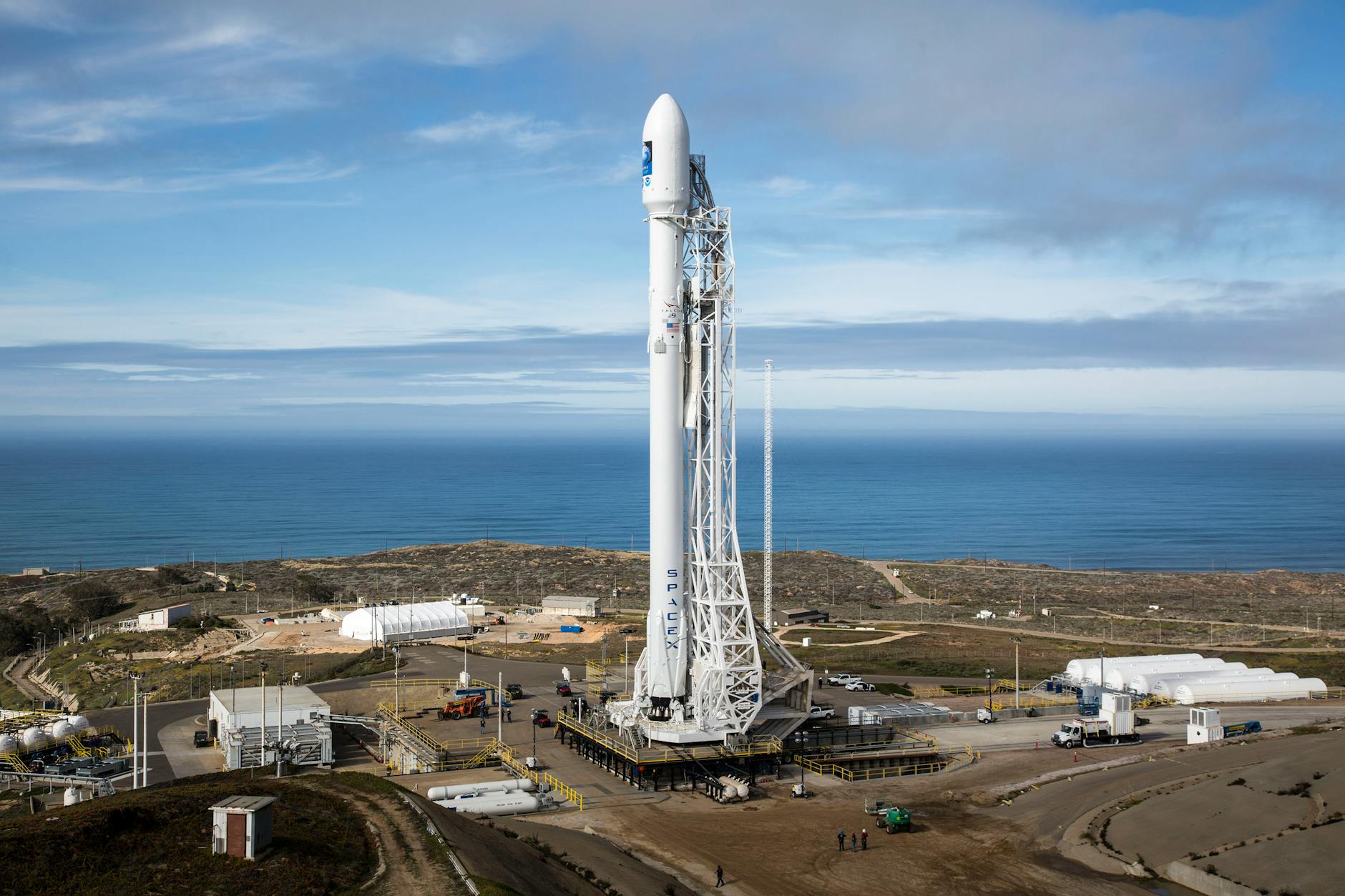 A SpaceX rocket stands on a launch pad overlooking the ocean under a clear sky.